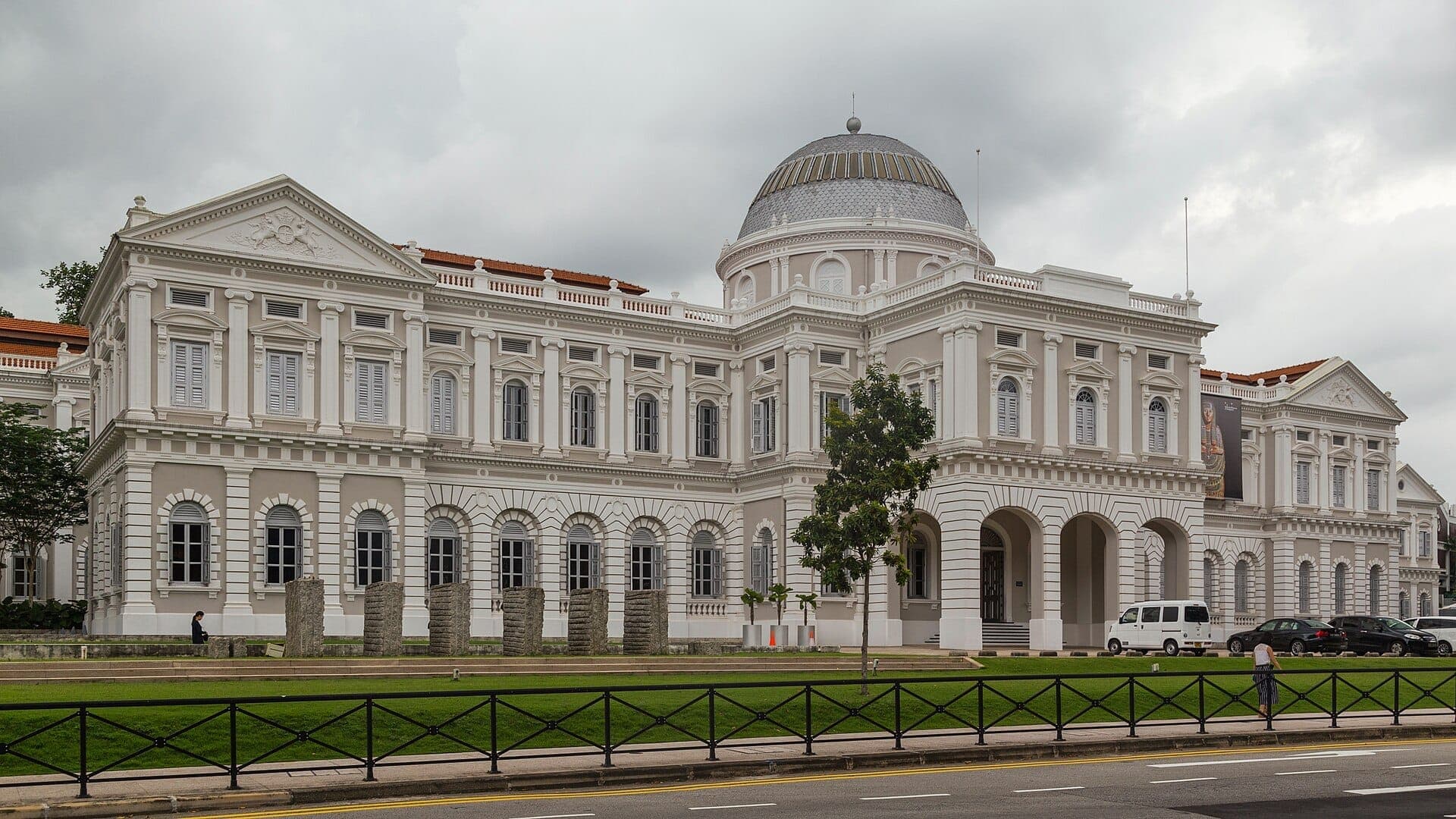 Wide view of the National Museum of Singapore’s grand colonial facade and central dome against a cloudy sky, with greenery and parked cars in front.
