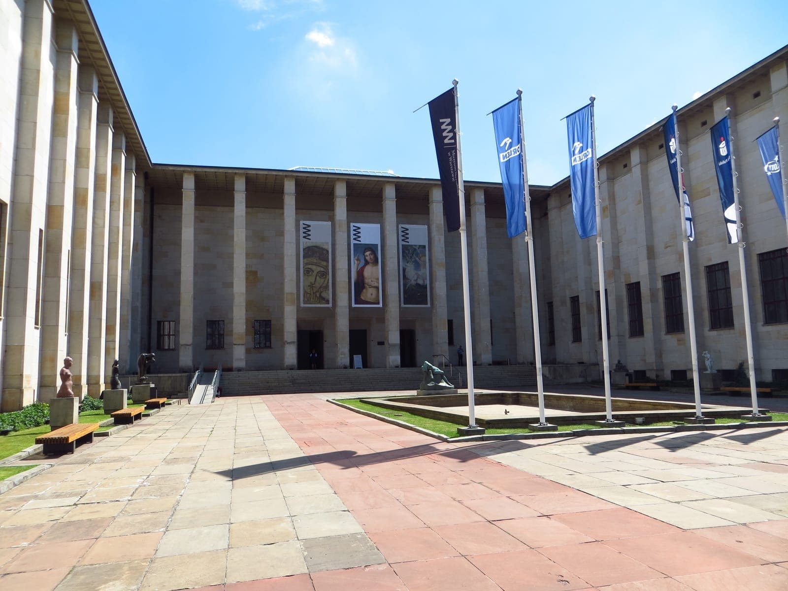 Der prächtige Eingang des Nationalmuseums in Warschau, mit Säulen, blauen Fahnen, Skulpturen im Freien und Kunstbannern an einem sonnigen Tag.