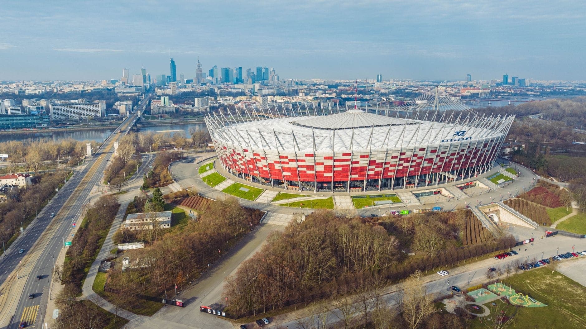 Luftaufnahme des PGE Narodowy am Ostufer der Weichsel mit der Warschauer Skyline, Bäumen, Brücken und Straßen rund um die moderne rot-weiße Arena.