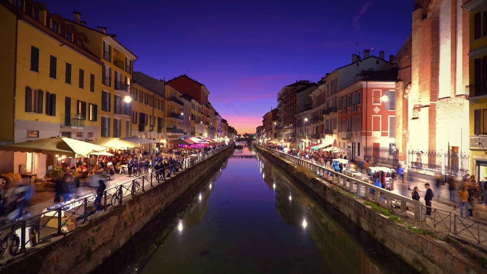 Evening view of the Navigli canal in Milan with vibrant lights, colorful buildings, reflections on the water, and lively crowds along both sides of the canal.