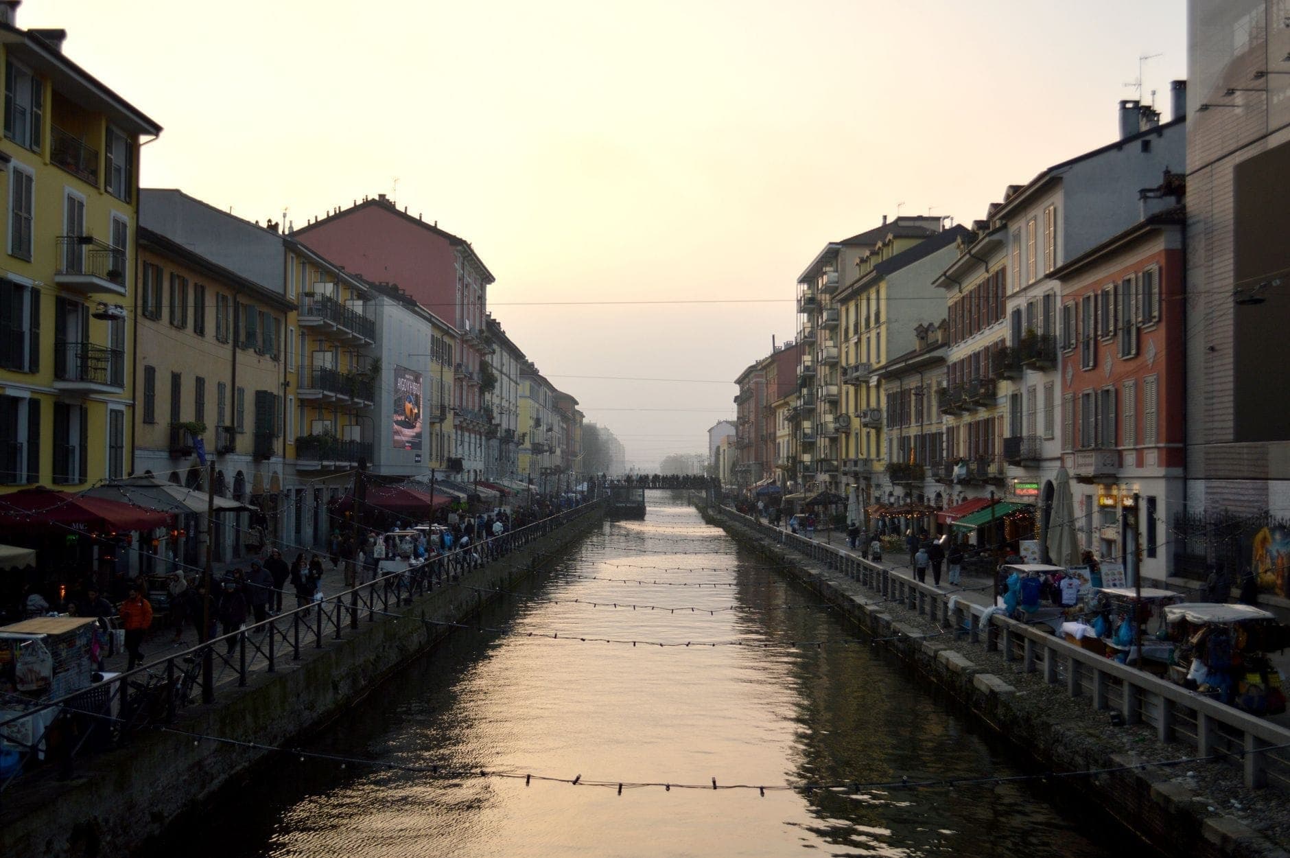 Evening view along Milan’s Naviglio Grande canal with colorful historic buildings, bustling crowds and reflections in the water under a glowing sunset sky.