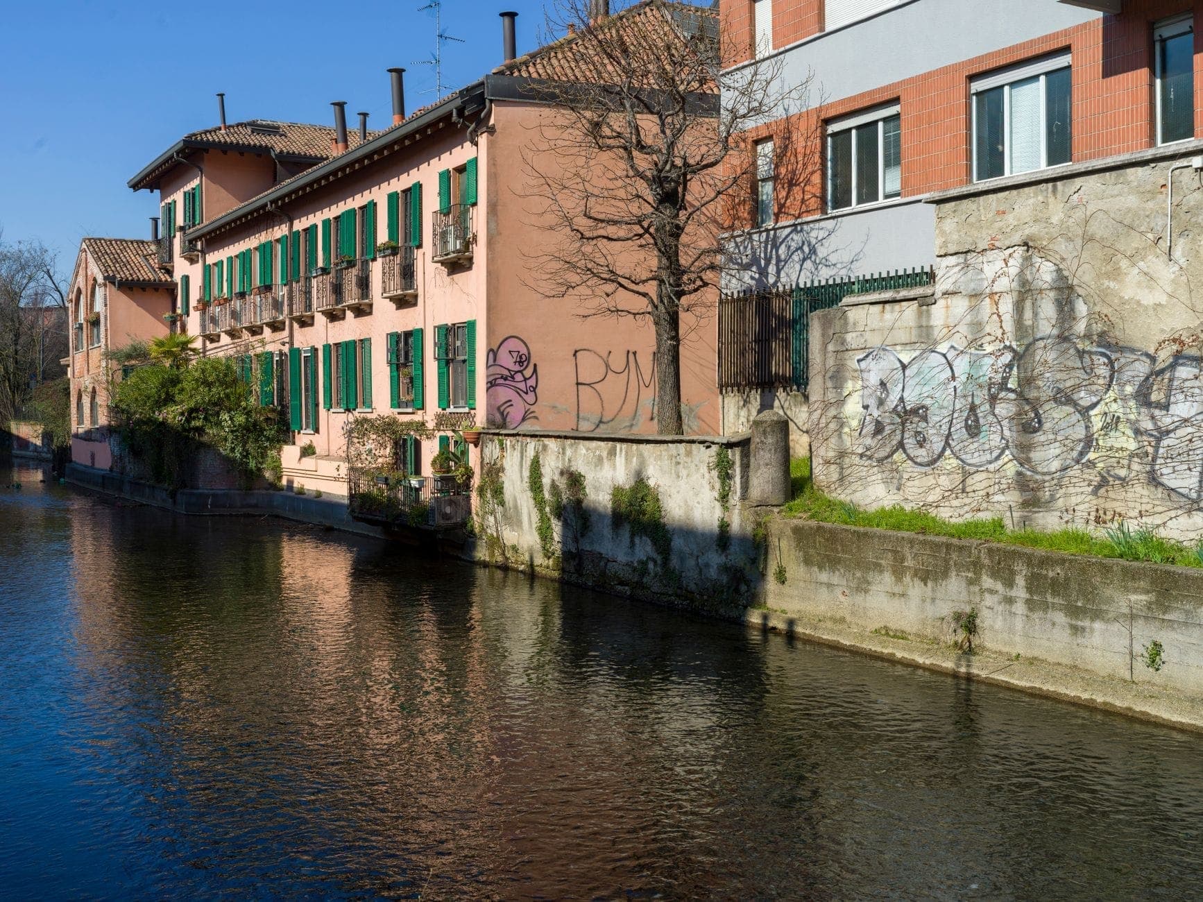 Calm canal scene showing traditional pink and brick buildings with green shutters, tree reflections in water, and peaceful atmosphere along the Naviglio Pavese in Milan.