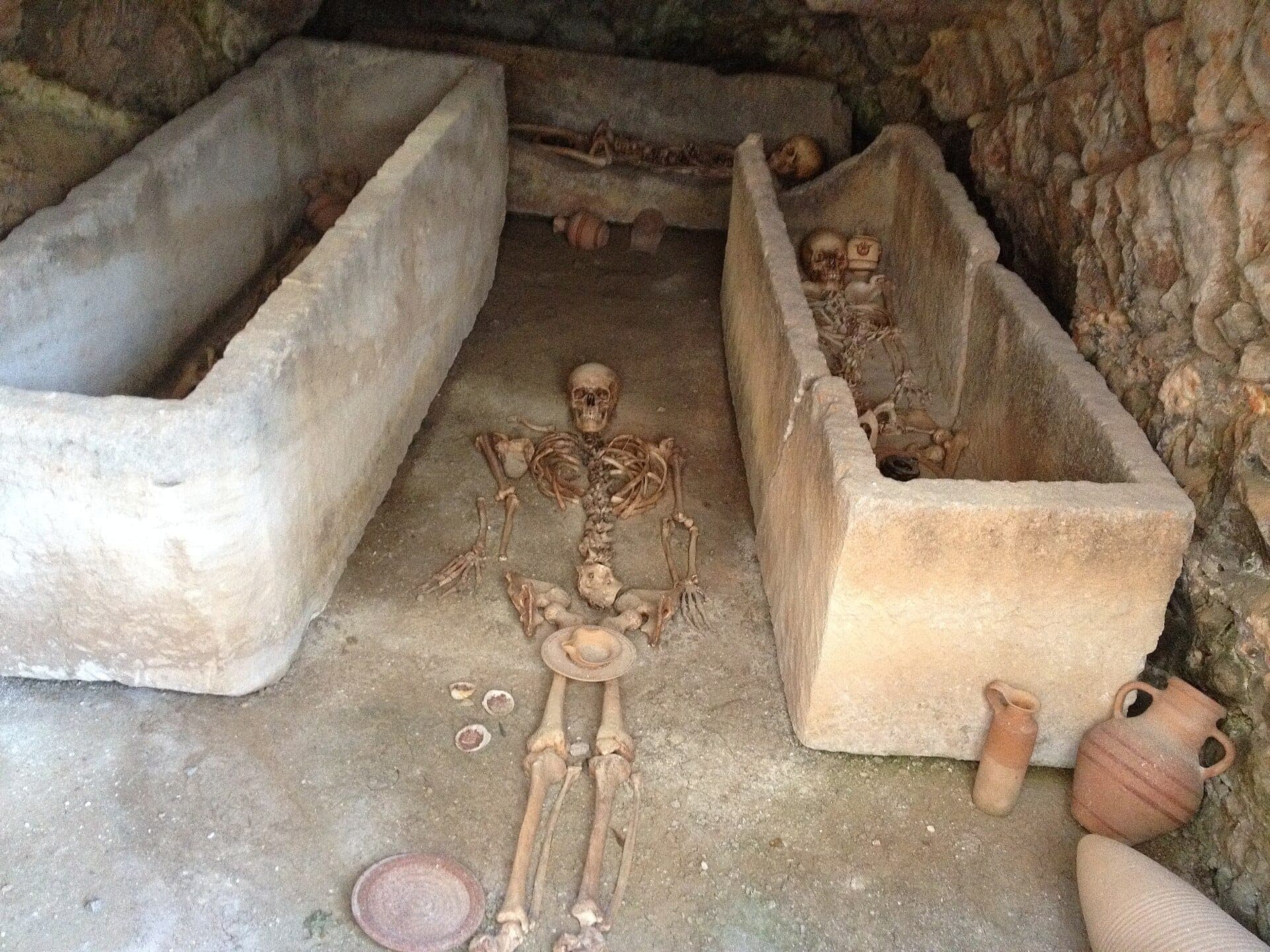 Ancient stone tombs with human skeletons and pottery at Necropolis del Puig des Molins in Ibiza, surrounded by rough stone walls.