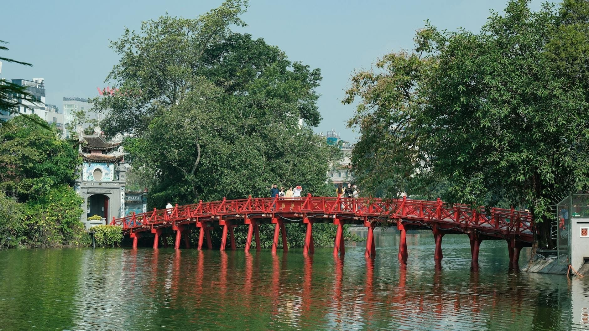 Toàn cảnh cầu Thê Húc sơn đỏ dẫn vào Đền Ngọc Sơn trên hòn đảo xanh mướt giữa Hồ Hoàn Kiếm Hà Nội, với du khách đang qua cầu.