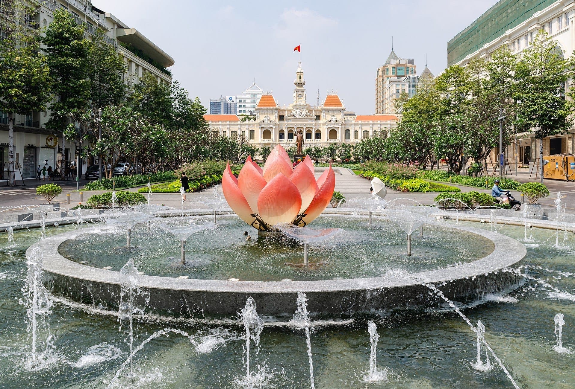 Đài phun nước hoa sen màu hồng lớn được bao quanh bởi các tia nước ở trung tâm Đường đi bộ Nguyễn Huệ, với Trụ sở Ủy ban Nhân dân ở phía sau.