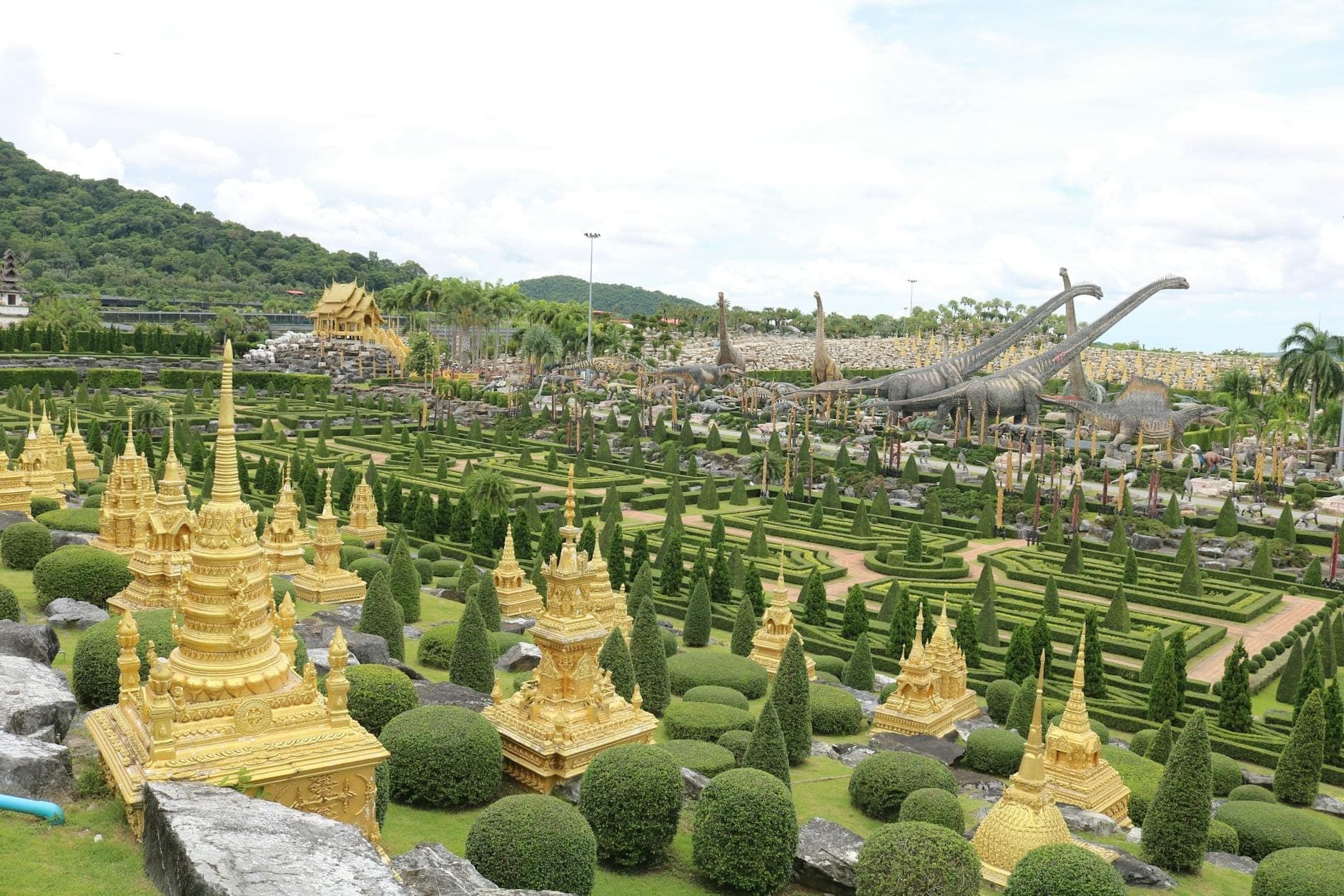 Wide view of Nong Nooch Tropical Garden with golden stupa structures, neatly manicured hedges, topiary trees, and distant dinosaur statues under a partly cloudy sky.
