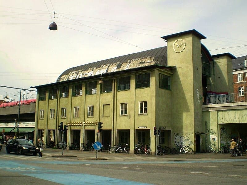 Straßenansicht der Nørrebro Station in Kopenhagen mit ihrer hellgelben Fassade, gewölbtem Dach, Stadtuhren, Graffiti, Fahrrädern und Fußgängern unter bewölktem Himmel.