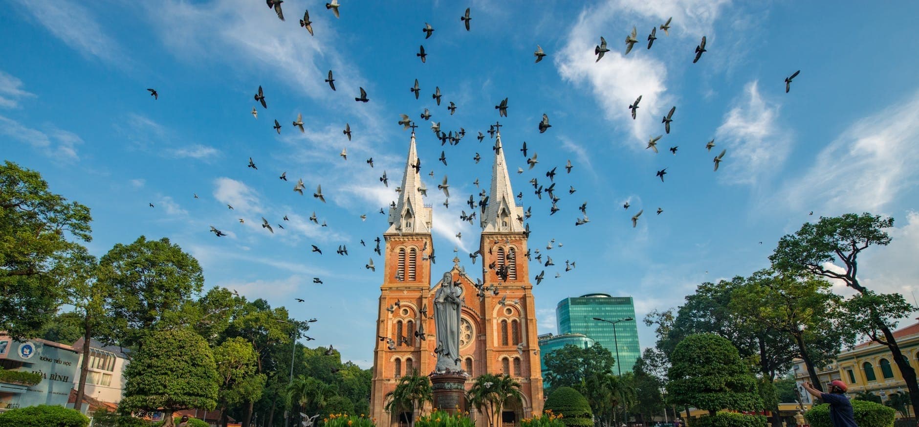 Nhà thờ Đức Bà Sài Gòn với hai tháp chuông gạch đỏ vươn cao, bầu trời xanh và đàn chim bay lượn phía trên tượng Đức Mẹ.