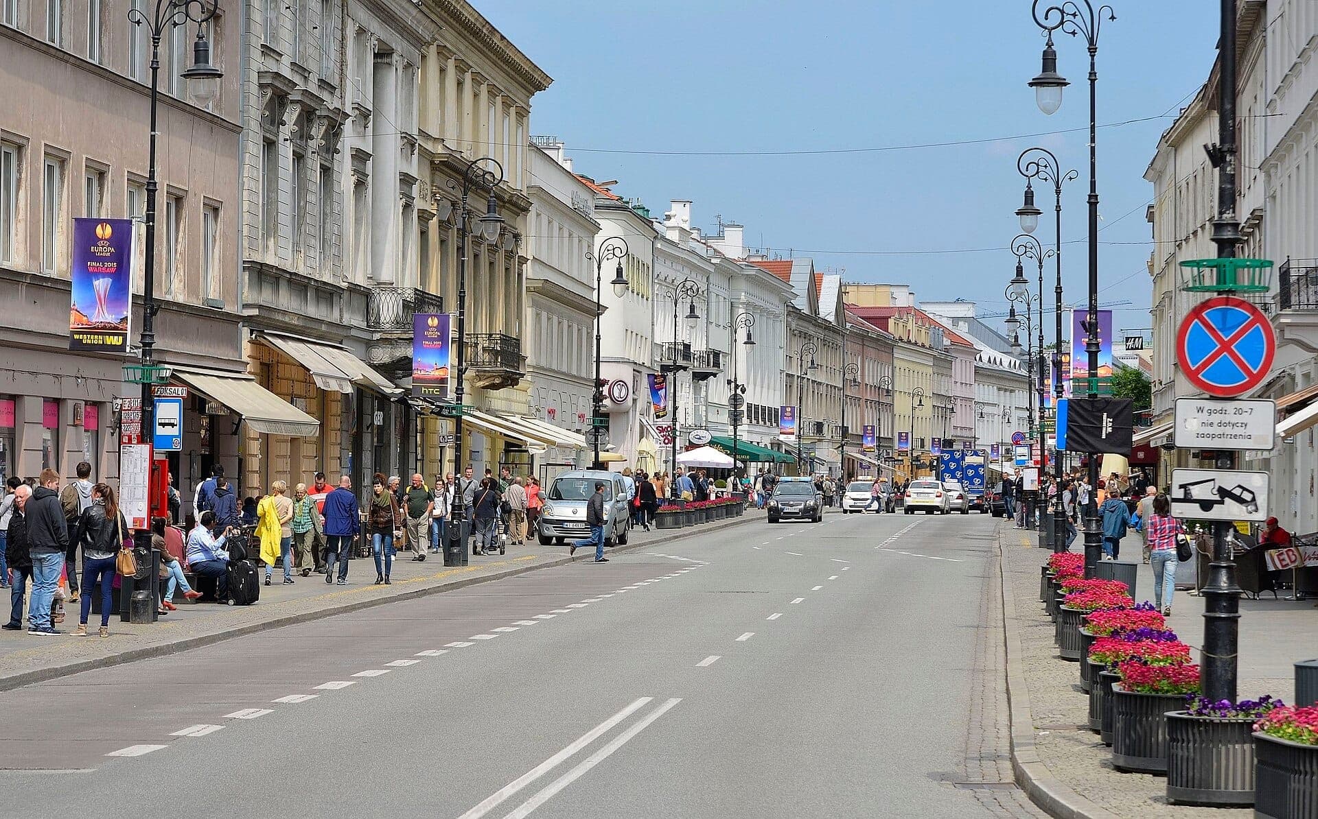 Die belebte Nowy Świat Straße in Warschau zeigt neoklassizistische Gebäude, Straßencafés, Straßenlaternen und Menschen, die an bunten Blumendekorationen entlanggehen, an einem hellen Tag.
