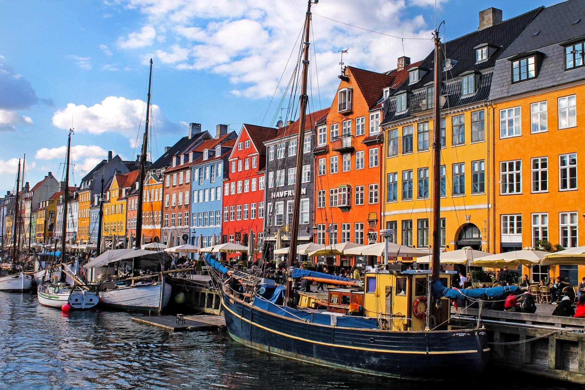 Bunt bemalte Nyhavn-Stadthäuser und historische Holzschiffe entlang des Kanals an einem sonnigen Tag, mit Menschen beim Essen im Freien.