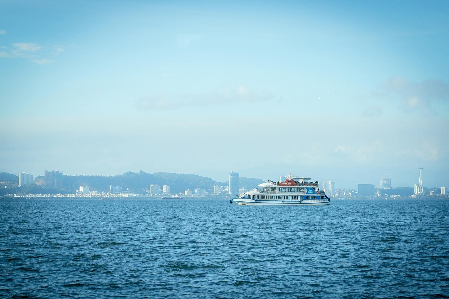 A multi-deck yacht cruises on the calm blue waters of Pattaya Bay with the city skyline and hills visible in the background under a clear sky.