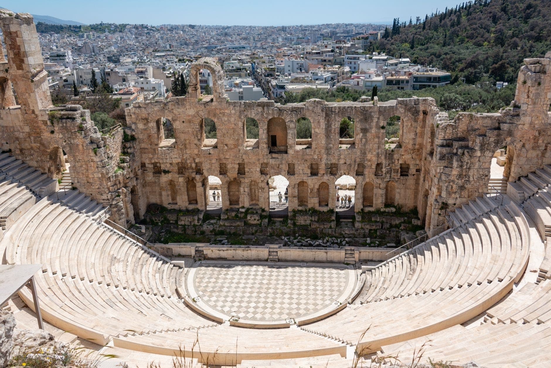 Wide-angle view of the Odeon of Herodes Atticus showing stone theater seats, arched stage wall, and Athens cityscape under bright daylight.