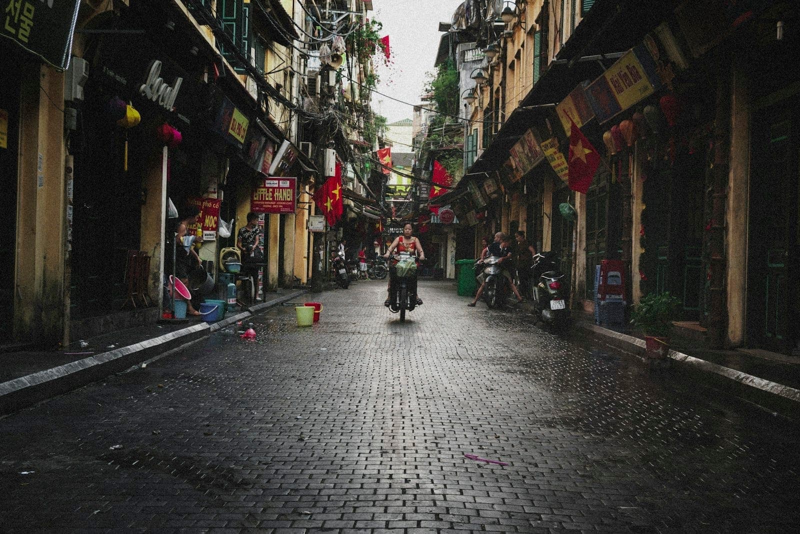 Isang makipot na kalye sa Old Quarter ng Hanoi, napapalibutan ng mga lumang kolonyal na gusali, mga watawat ng Vietnam, at mga parol, habang may isang tao na nagbibisikleta sa makitid na basa-basang daan.