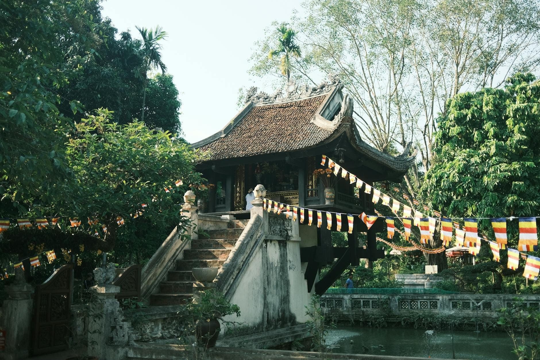 Chùa Một Cột — ngôi chùa gỗ nhỏ nhô lên trên một trụ đá giữa hồ sen vuông, xung quanh là cây xanh và cờ Phật giáo nhiều màu sắc tại Hà Nội, Việt Nam.