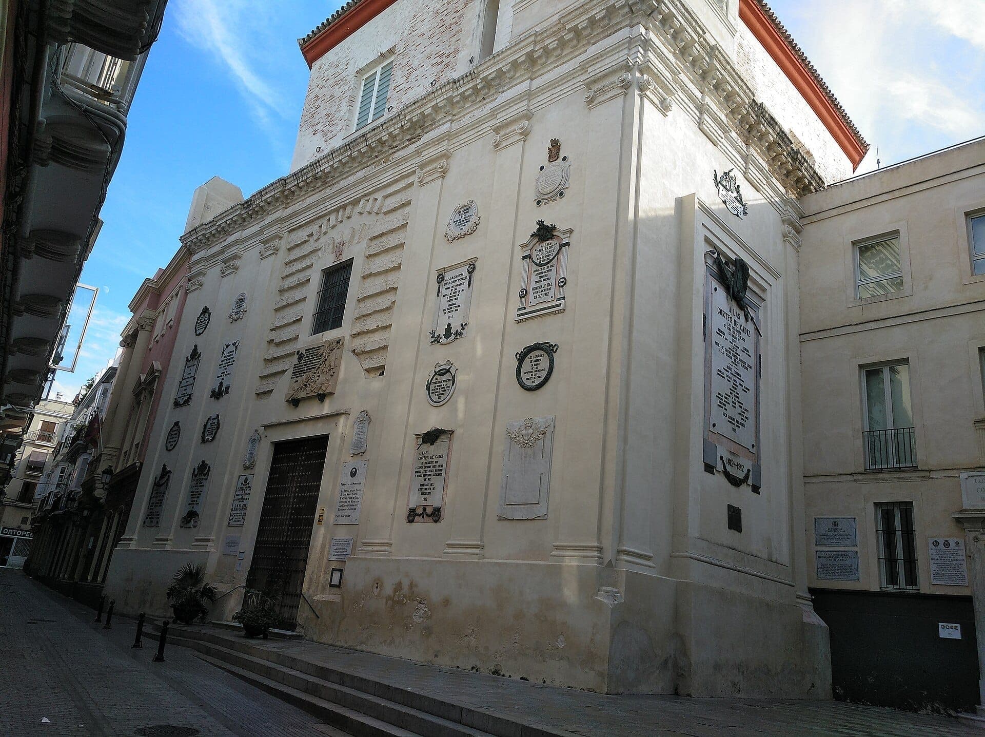 Mặt tiền của Oratorio de San Felipe Neri tại Cádiz, nổi bật với những bức tường Baroque màu kem được trang trí bằng nhiều tấm biển kỷ niệm dưới bầu trời xanh trong.