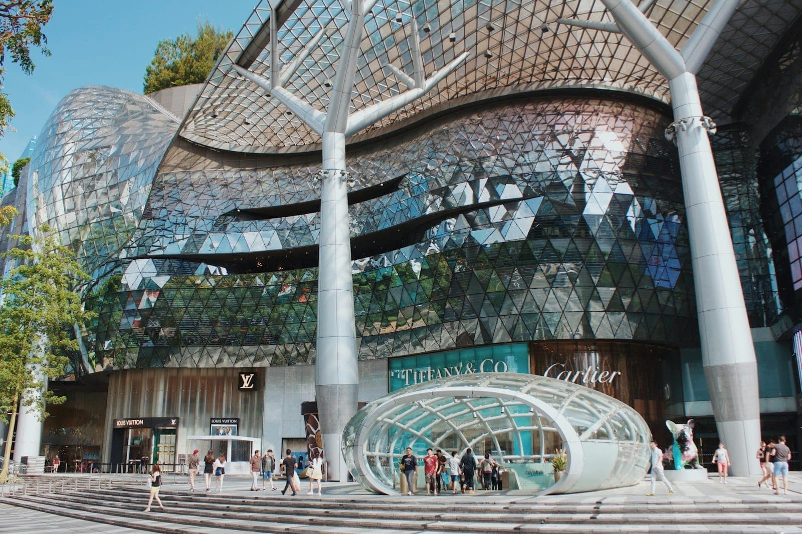 Exterior of a modern glass-front shopping mall on Orchard Road, featuring luxury brand storefronts and people walking past a futuristic entrance.