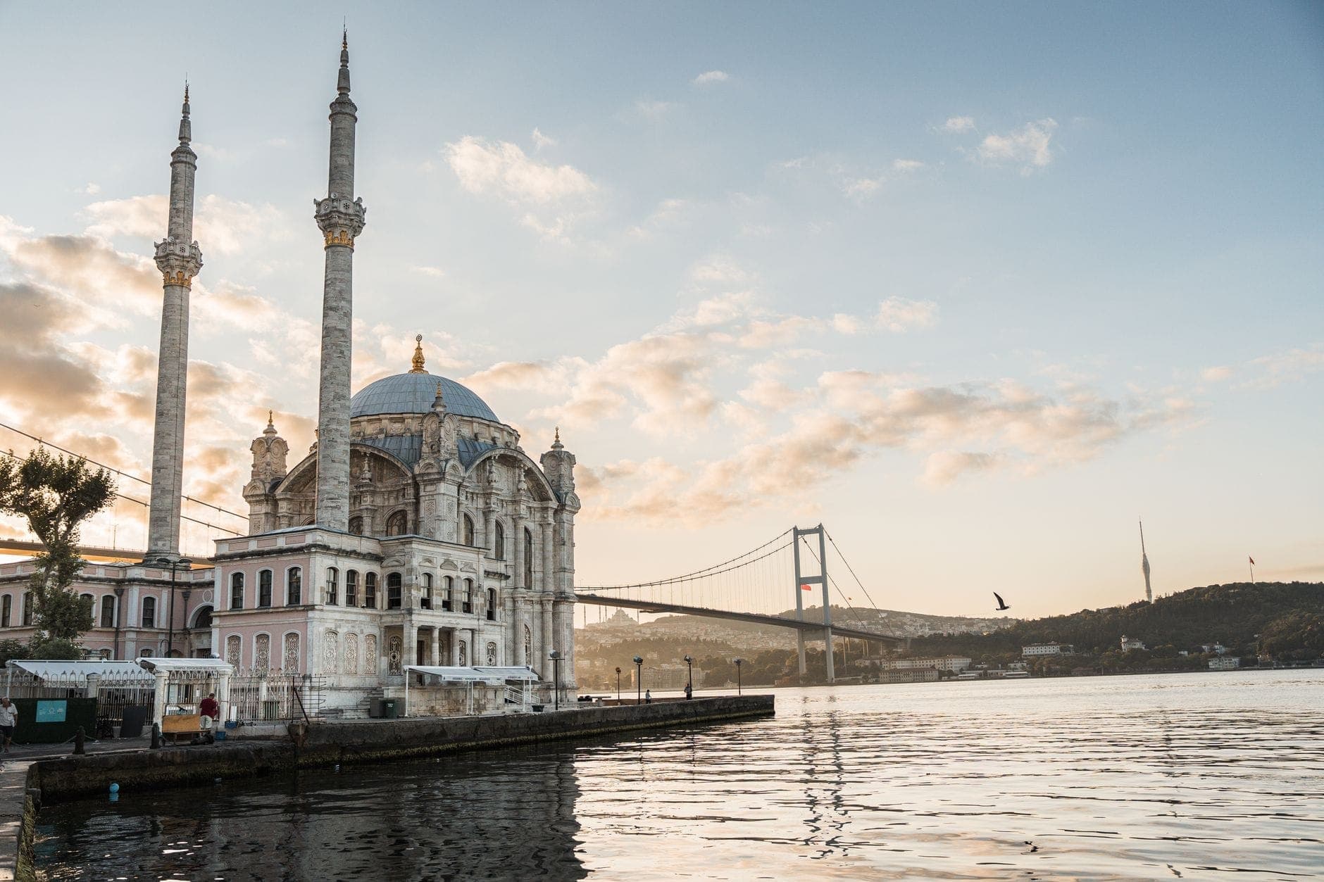 Die Ortaköy-Moschee bei Sonnenaufgang am Bosporus-Ufer, mit der Bosporus-Brücke im Hintergrund und goldenem Licht, das sich im Wasser spiegelt.