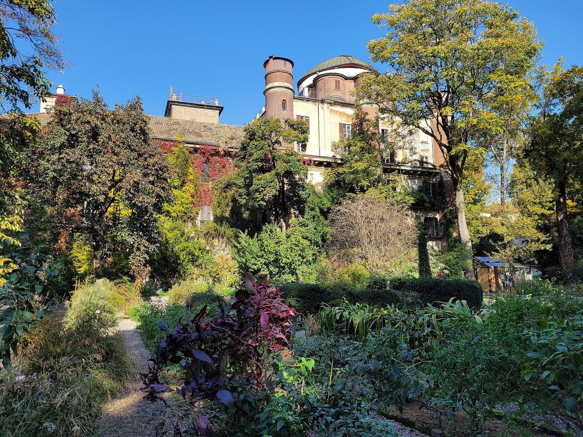 Sunlit view of lush gardens, mature trees, and the historic Palazzo Brera buildings in the Orto Botanico di Brera, Milan.