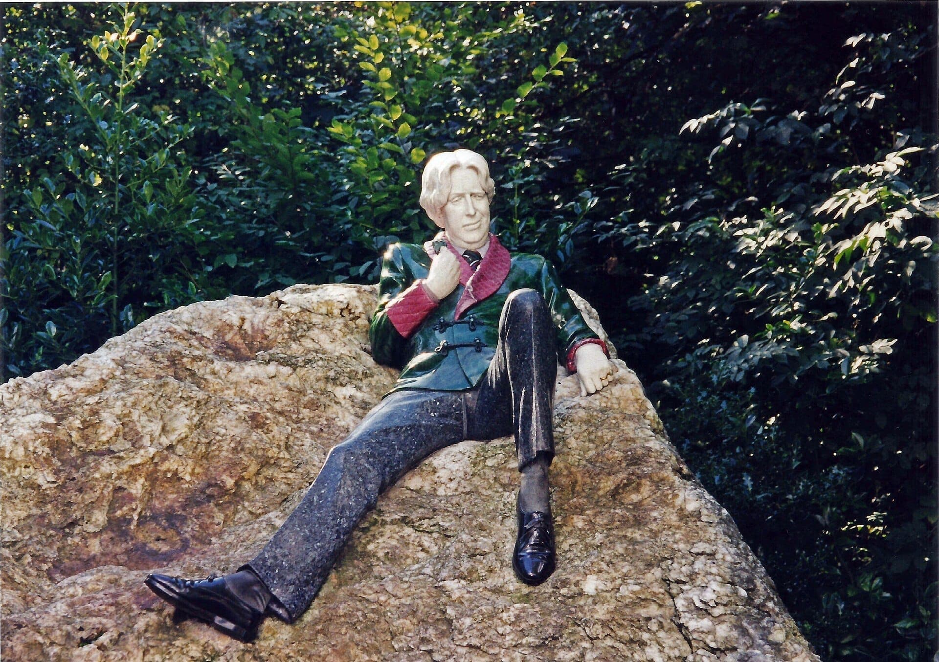 Escultura conmemorativa de Oscar Wilde recostada sobre una gran roca de cuarzo en Merrion Square, rodeada de exuberante follaje verde a la luz natural del día.