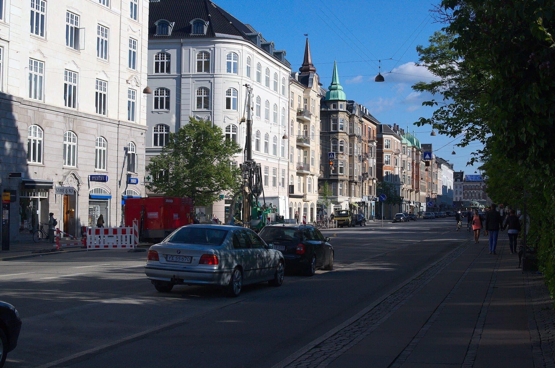 Baumgesäumter Boulevard in Østerbro, Kopenhagen, mit historischen mehrstöckigen Gebäuden, am Straßenrand geparkten Autos und einem klaren blauen Himmel.