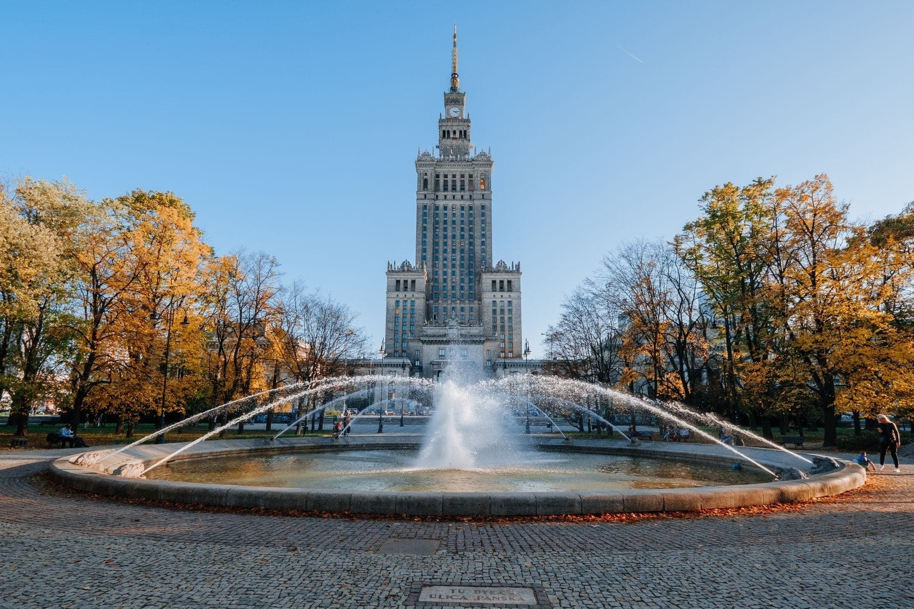 Der Palast der Kultur und Wissenschaft ragt über einen großen Springbrunnen mit herbstlichen Bäumen in Warschau empor, aufgenommen von einem zentralen Weg unter einem klaren, hellen Himmel.