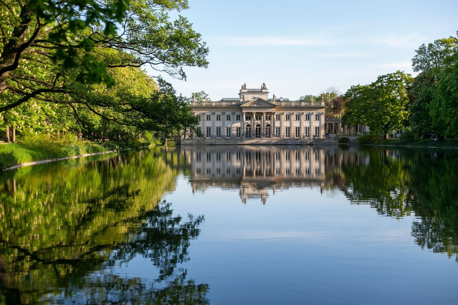 Blick auf den Palast auf der Insel in Warschau, der sich perfekt in einem stillen See spiegelt, umgeben von üppig grünen Bäumen unter strahlend blauem Himmel.