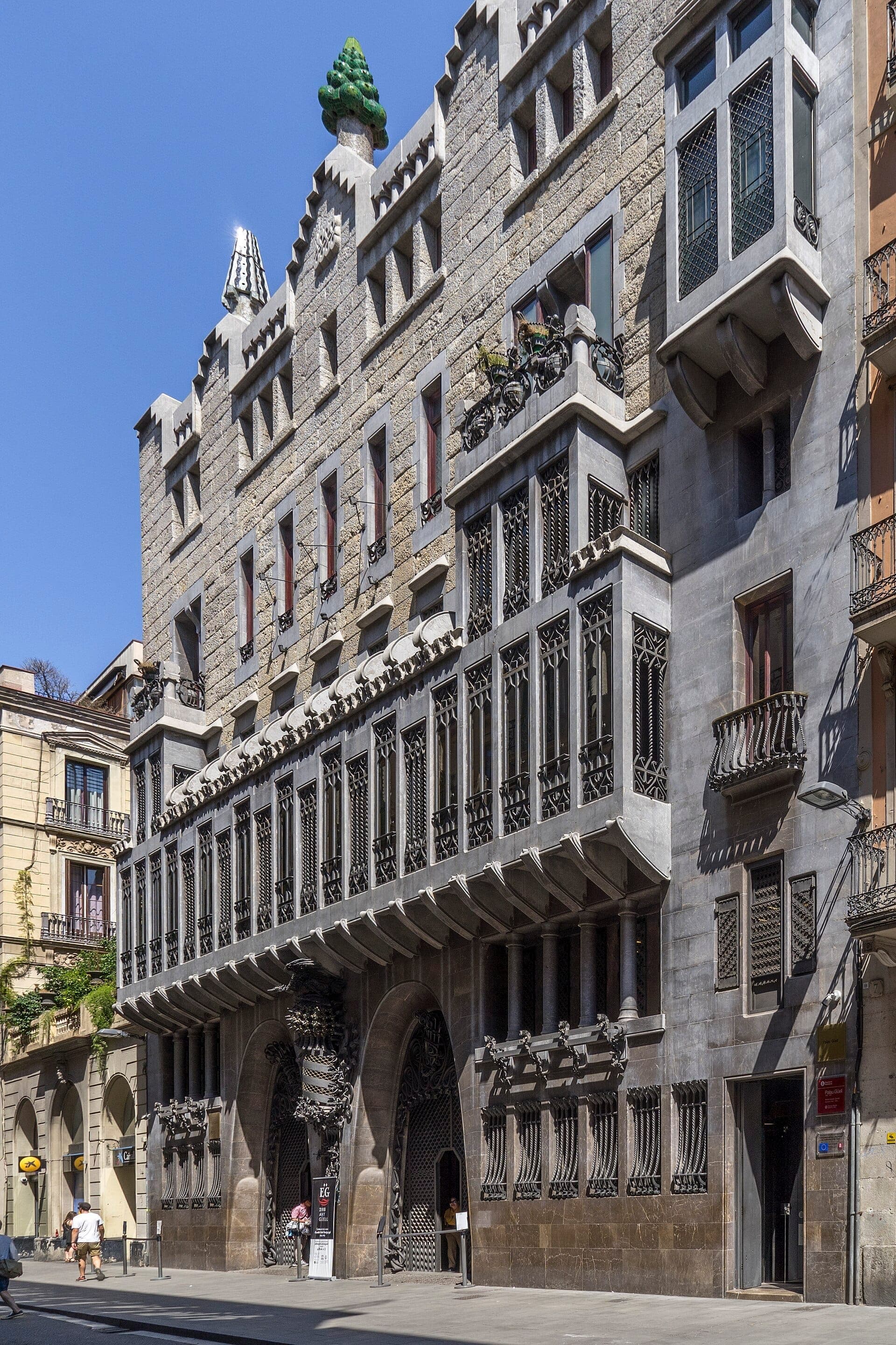 Mặt tiền của Palau Güell ở Barcelona, nổi bật với các chi tiết sắt rèn tinh xảo, hình dạng cửa sổ độc đáo và các ống khói mái nhà đầy màu sắc dưới bầu trời xanh trong.