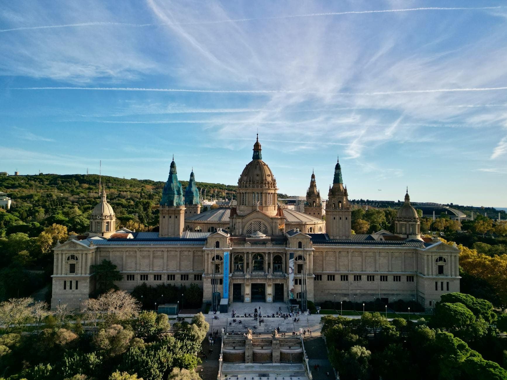 Toàn cảnh Palau Nacional trên đỉnh đồi Montjuïc nhìn từ trên cao, bao quanh là cây xanh, bầu trời xanh và những đám mây nhẹ, tại Barcelona.