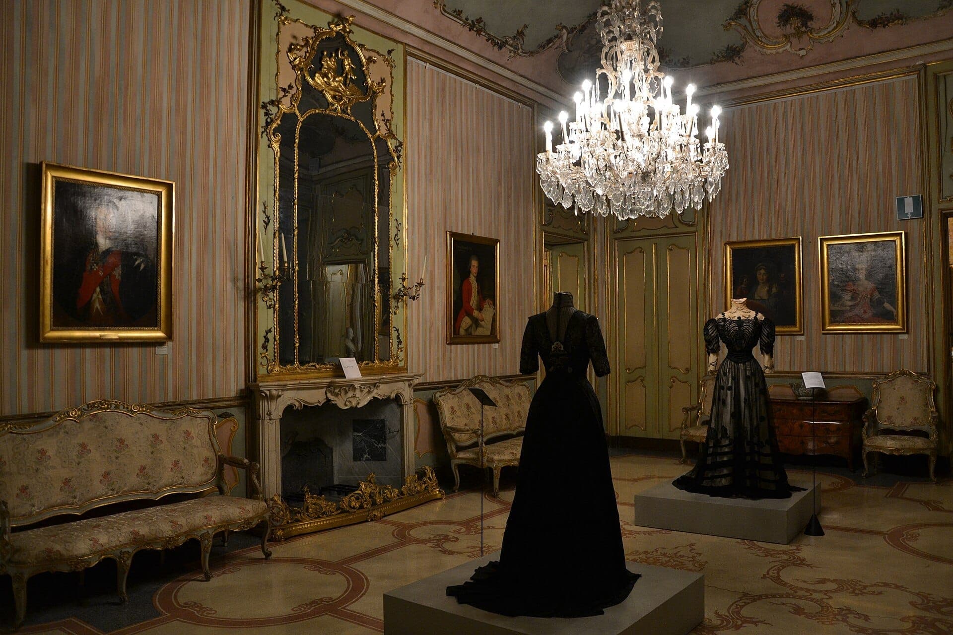 Two ornate black dresses on display in an opulent salon at Palazzo Morando, with antique sofas, gilded mirror, paintings, and a sparkling chandelier.
