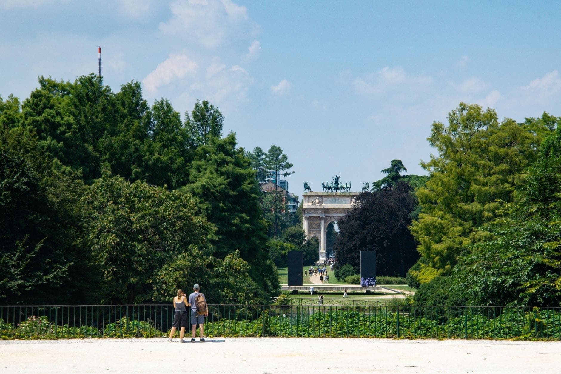 Wide view of Parco Sempione in Milan with lush greenery, people walking, and the iconic Arco della Pace visible in the distance under a blue sky.