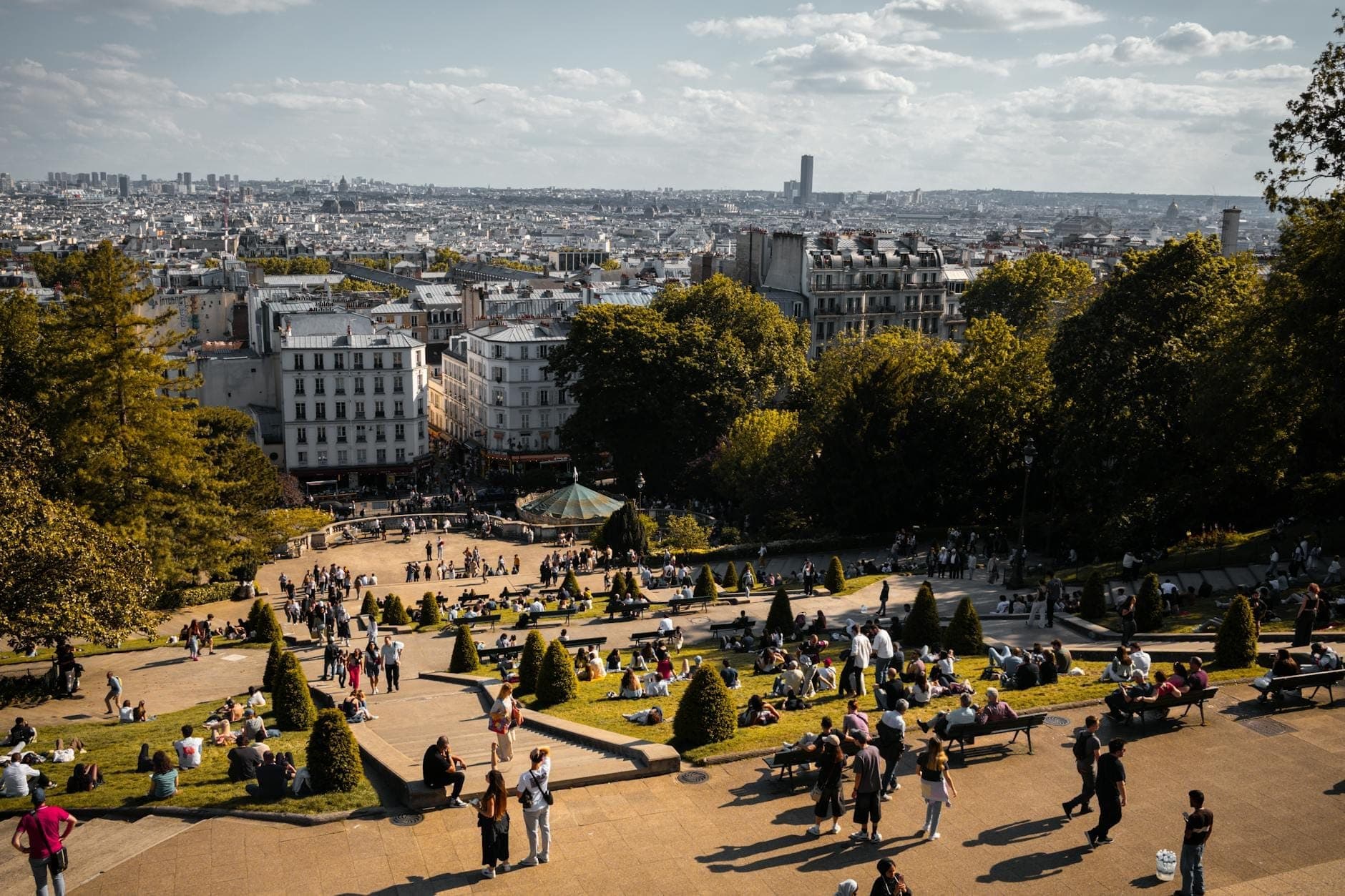 Toàn cảnh một công viên Paris nhộn nhịp với các gia đình và trẻ em, ghế băng, hàng cây và những tòa nhà cổ điển phía sau dưới bầu trời nắng đẹp.