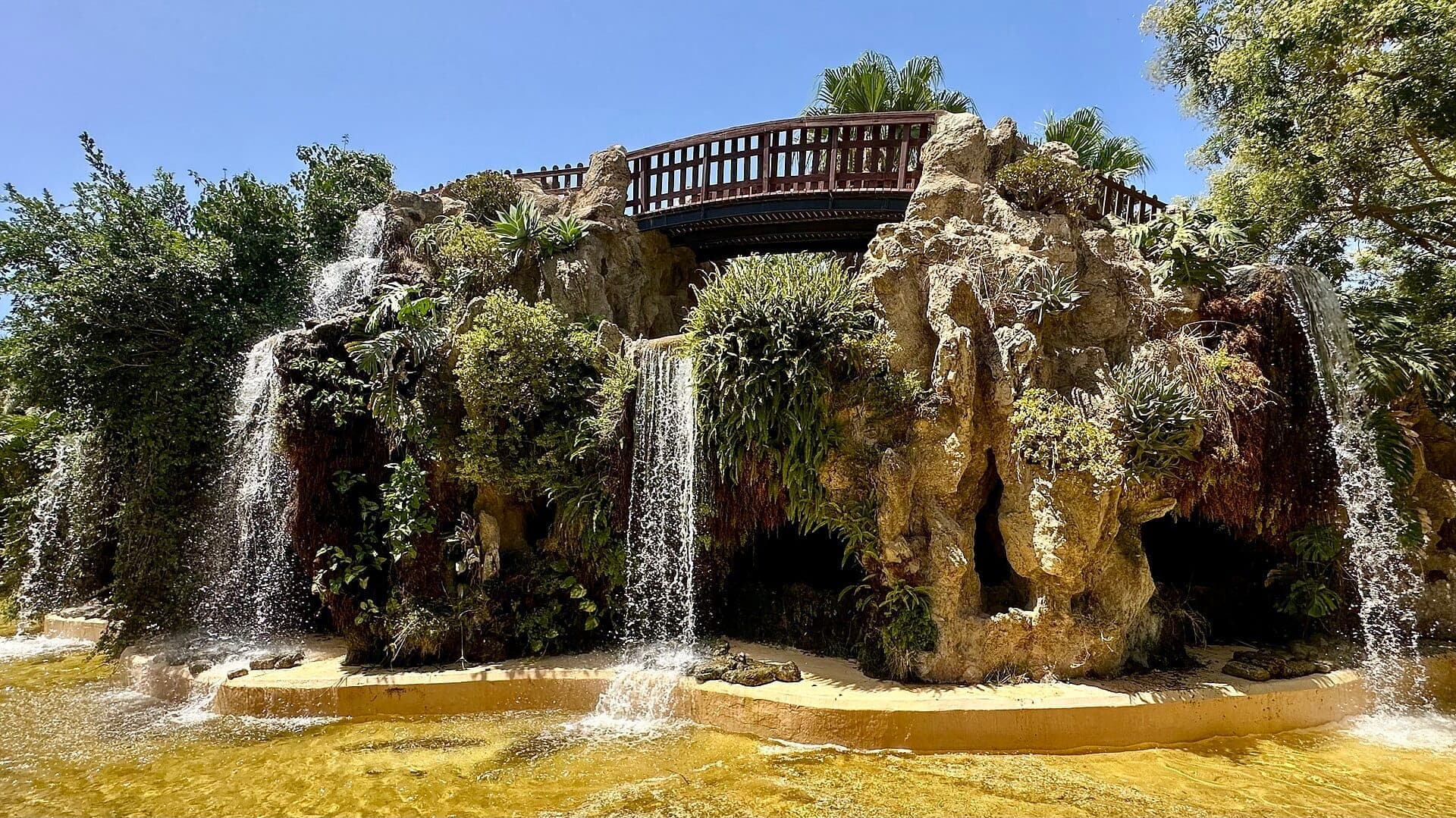 Cảnh thác nước tại Parque Genovés ngập nắng với thảm thực vật xanh tươi, các khối đá và cây cầu gỗ dưới bầu trời xanh trong tại Cádiz.