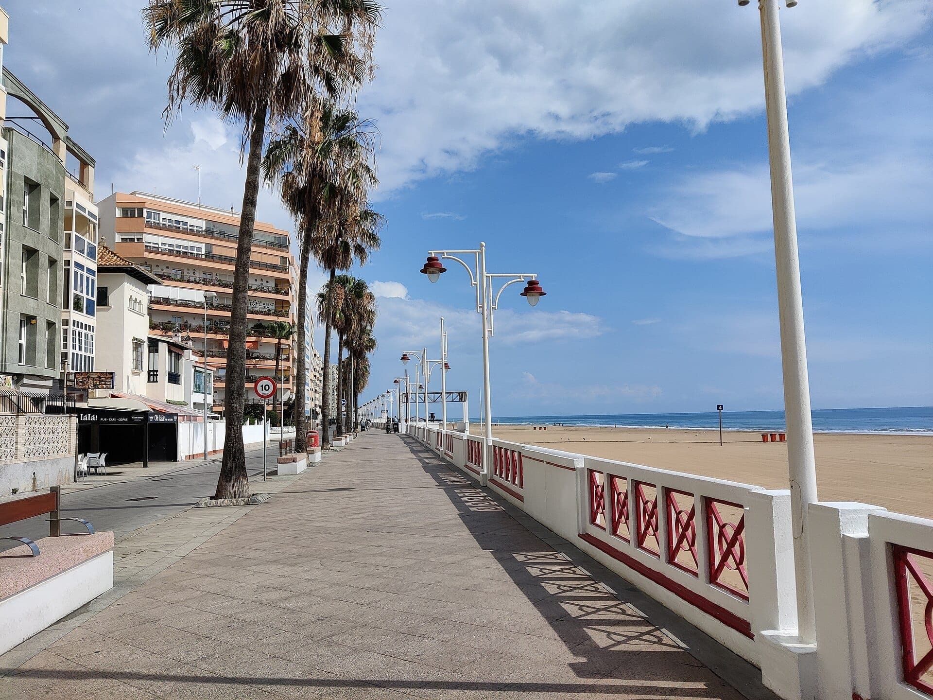 Paseo Marítimo de Cádiz rợp bóng cây cọ với những tòa nhà hiện đại ven biển, lan can trắng và tầm nhìn ra đại dương dưới bầu trời xanh trong sáng.
