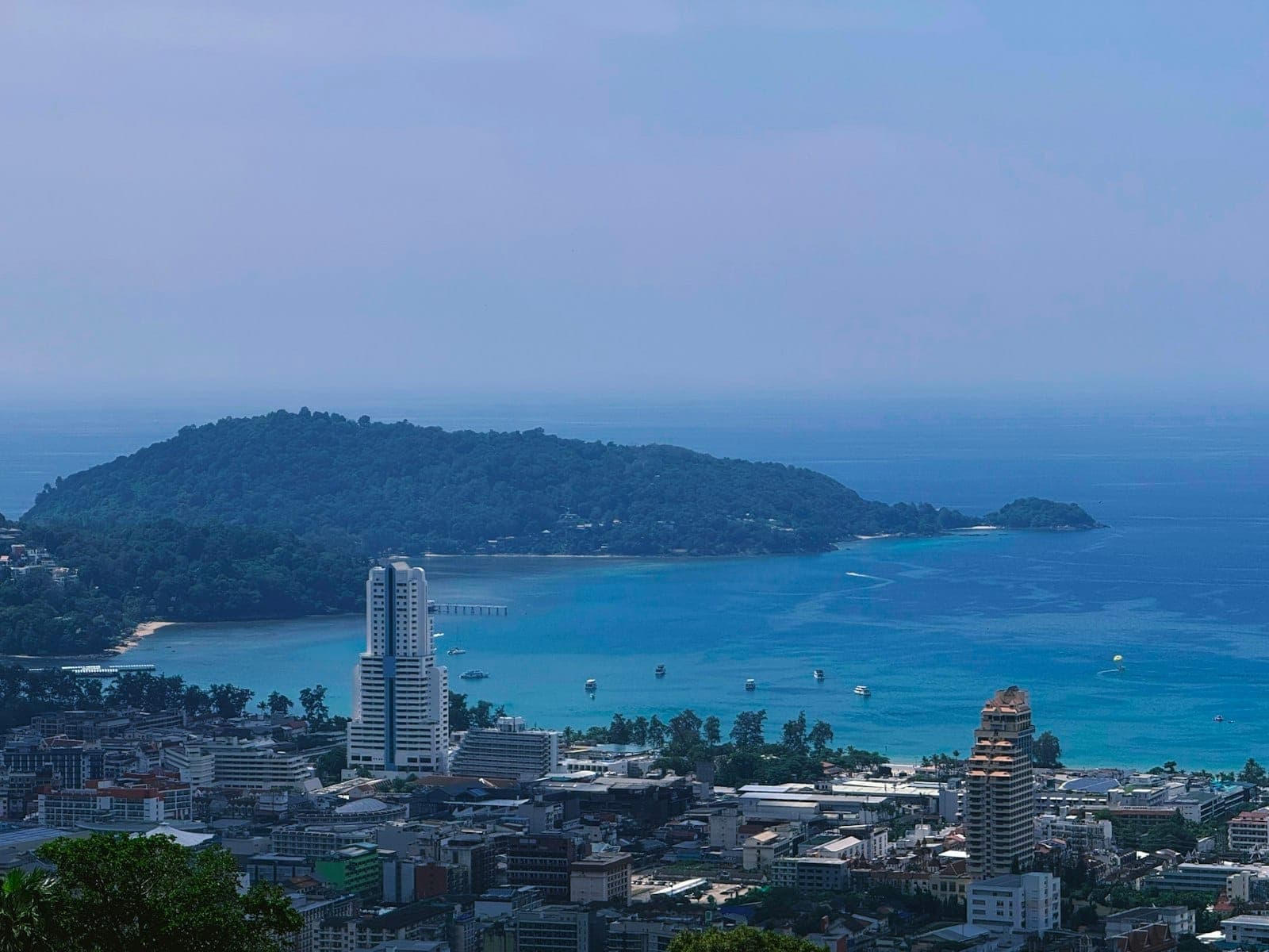 Panoramikong tingin sa look ng Patong na may turkesa-kulay na dagat, makabagong mataas na gusali, at makulay na lungsod na nakahanay sa tabi ng dalampasigan, sa ilalim ng maliwanag na asul na langit.
