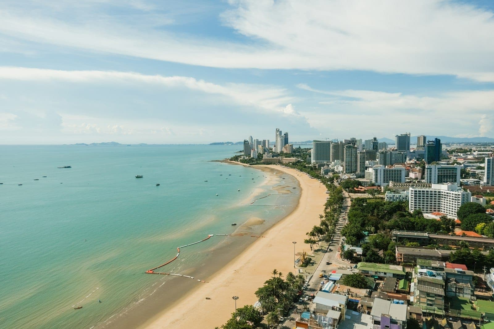 Aerial view of Pattaya Beach showing golden sand, turquoise water, beachfront high-rise hotels, and busy city streets under a bright, partly cloudy sky.