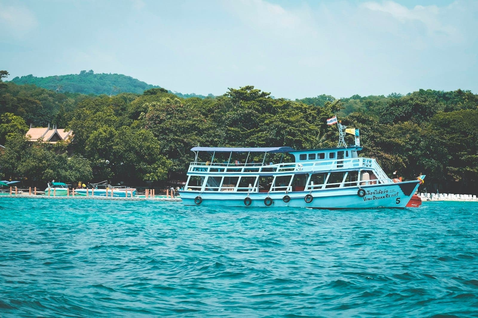 Turquoise ferry boat near the shore of a lush green island, with clear blue water and a partly cloudy sky.