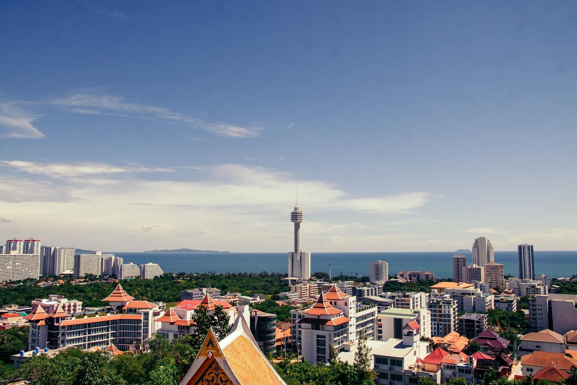 Wide cityscape view of Pattaya with hotels, skyscrapers, the recognizable Pattaya Tower, and the Gulf of Thailand coastline under a clear blue sky.