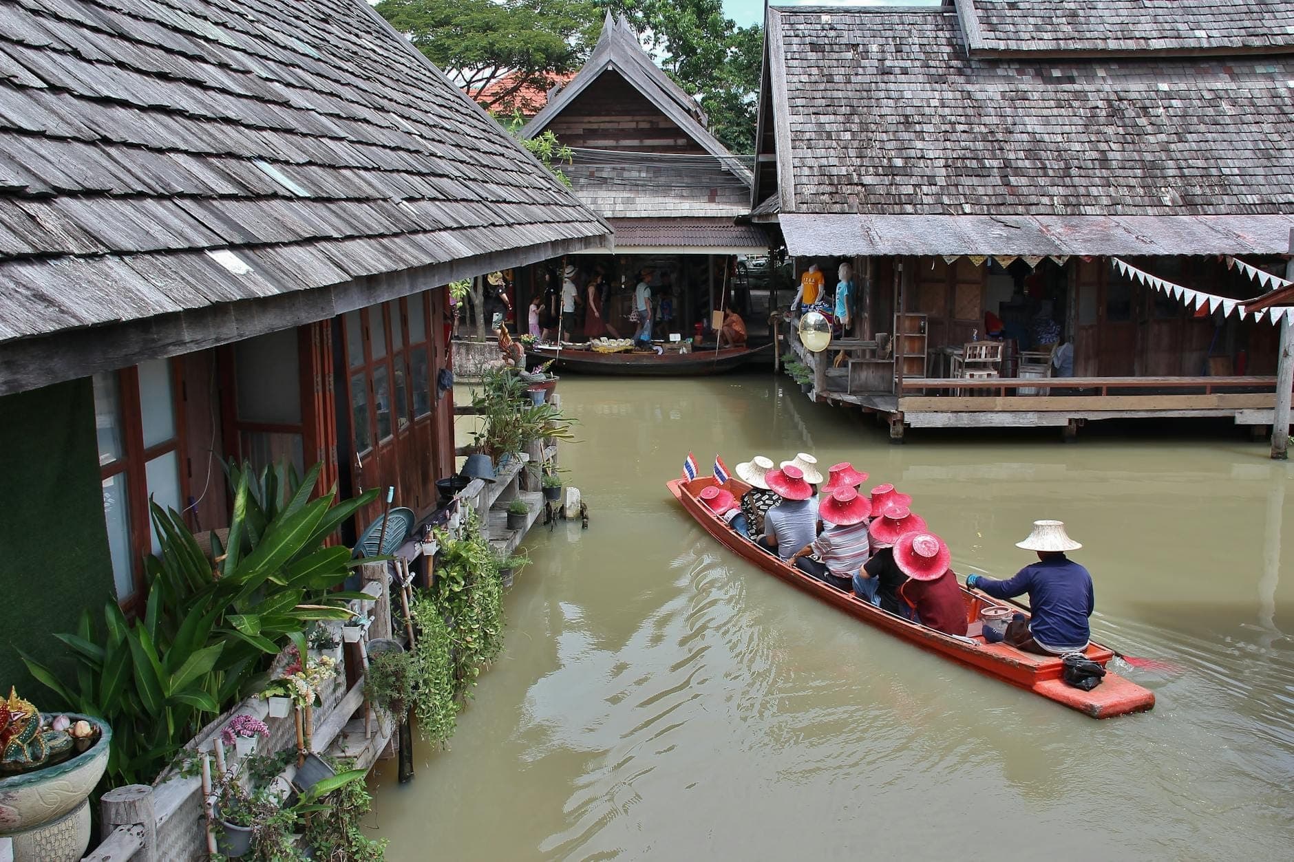 Wooden boats rowed by guides with tourists in colorful hats, gliding through narrow canals surrounded by rustic Thai stilt houses at Pattaya Floating Market.