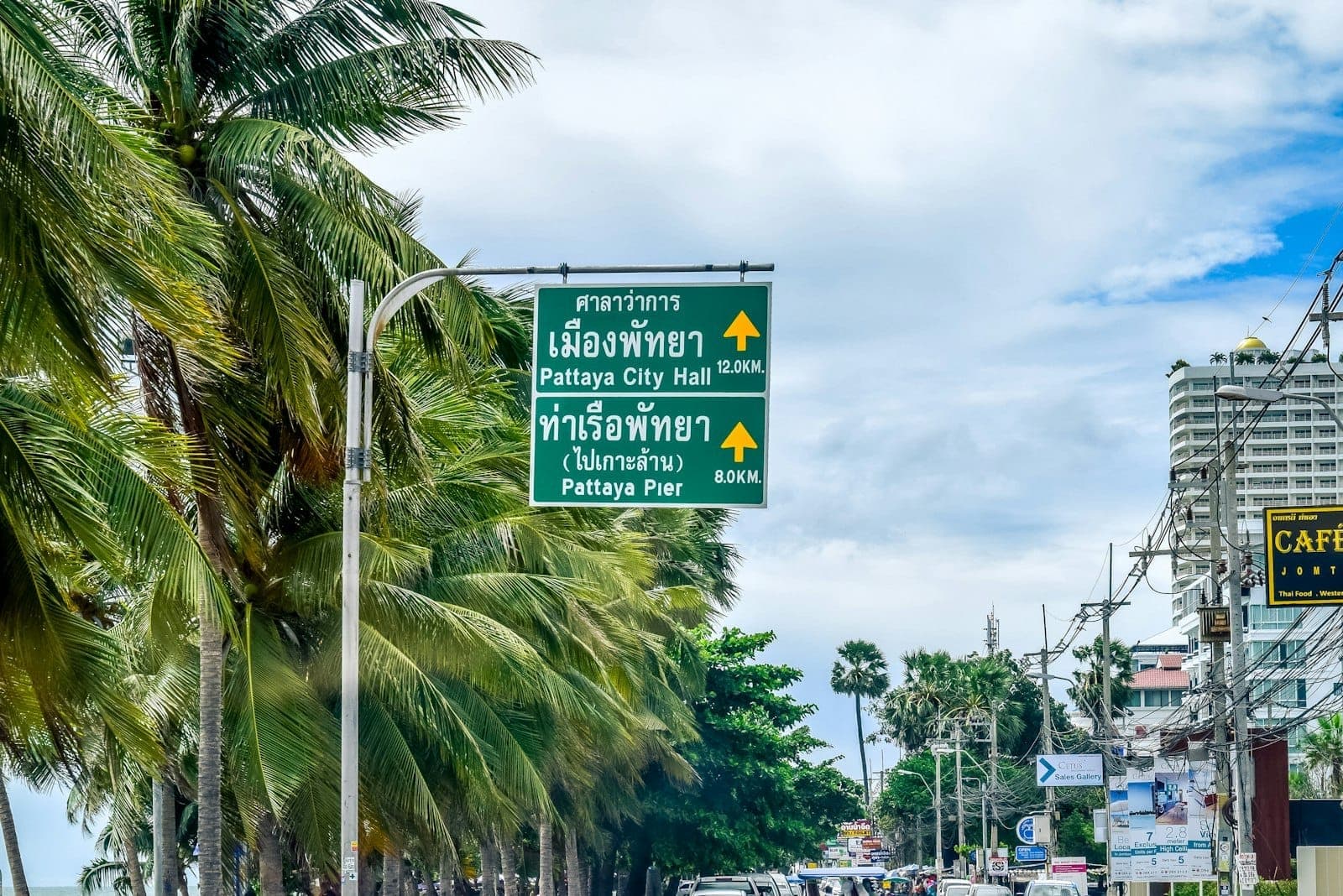 Green street signs for Pattaya City Hall and Pattaya Pier with palm trees, buildings, and traffic along a sunny coastal road.