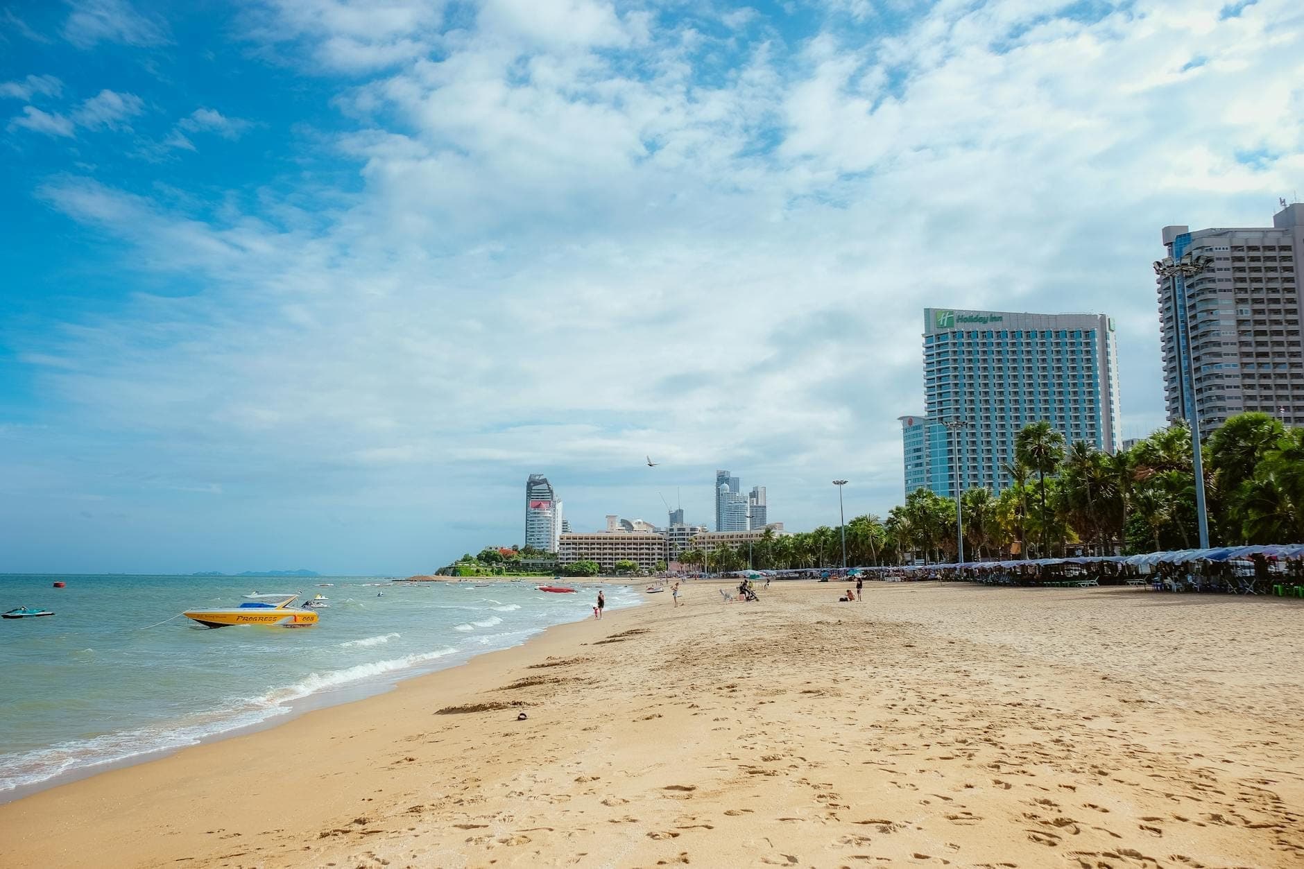 Wide view of Pattaya Beach with golden sand, gentle waves, yellow speedboat, palm trees, and city hotels lining the shoreline under a bright, partly cloudy sky.