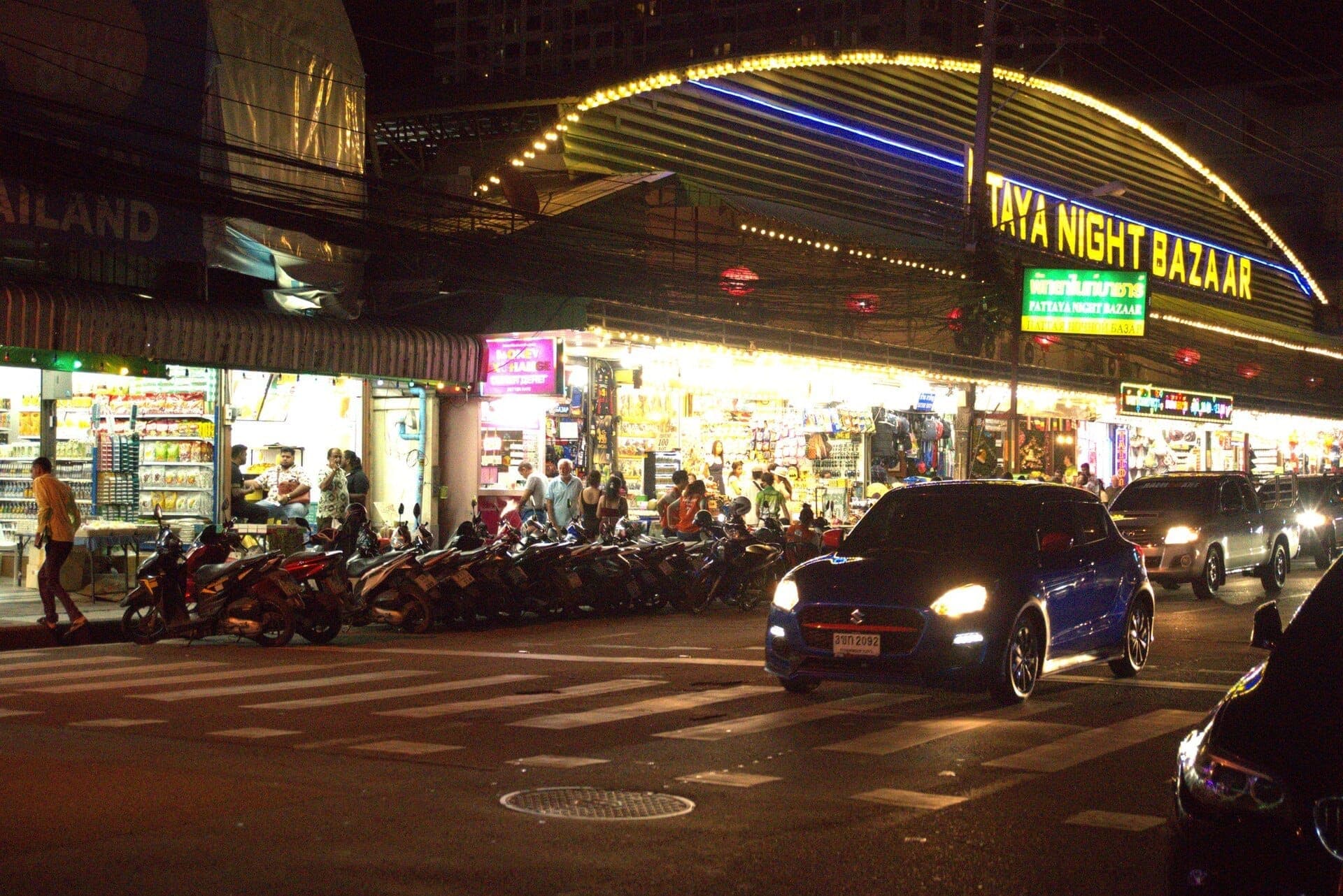 Bright lights illuminate the entrance to Pattaya Night Bazaar at night, with motorbikes and cars parked outside and shoppers visible inside.