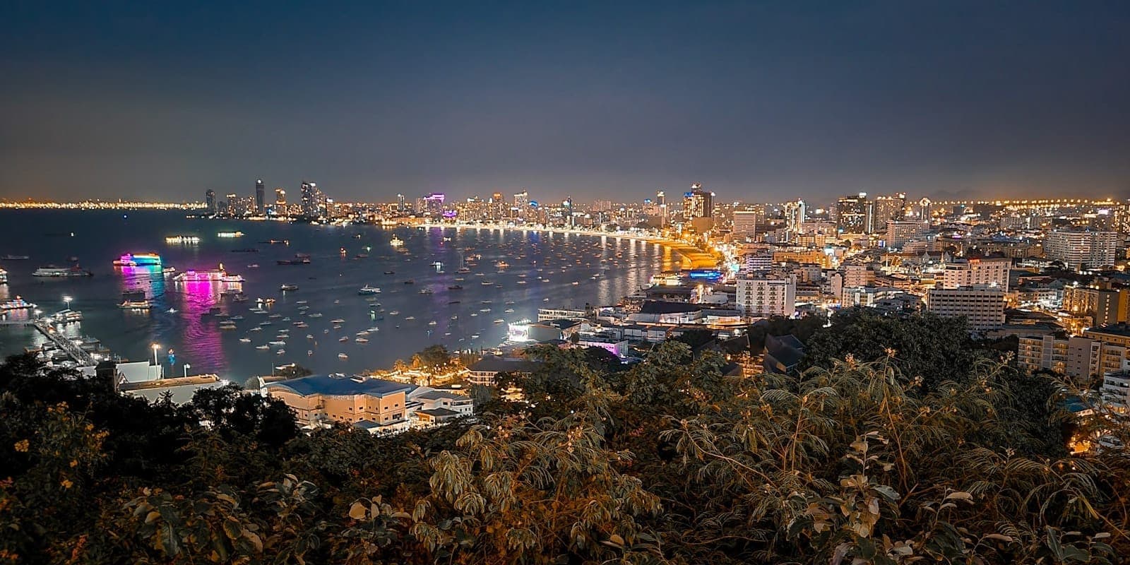Aerial night view of Pattaya city with illuminated coastline, boats in the bay, and colorful city lights.
