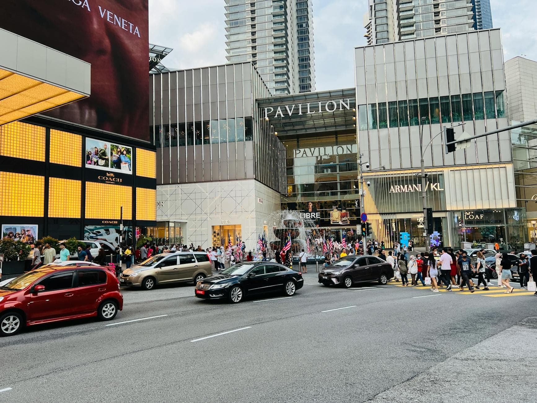 Cảnh đường phố tại lối vào chính của Pavilion Kuala Lumpur với các cửa hàng thương hiệu cao cấp, vạch sang đường đông đúc, các tòa nhà hiện đại và đông đảo người mua sắm.