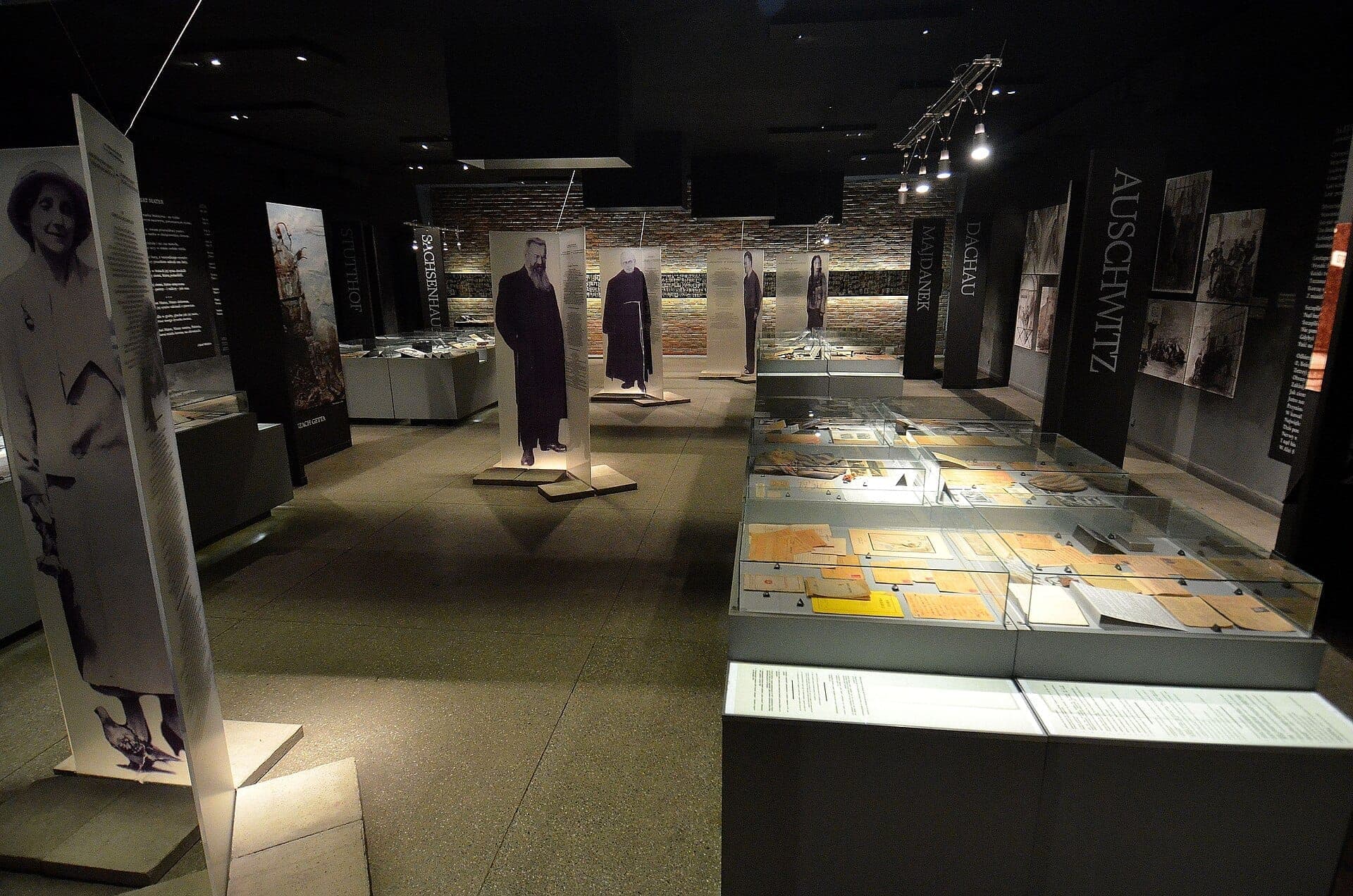 Innenraum des Pawiak-Gefängnismuseums mit historischen Ausstellungen, Glasvitrinen mit Dokumenten und lebensgroßen Porträts ehemaliger Gefangener bei gedämpfter Beleuchtung.