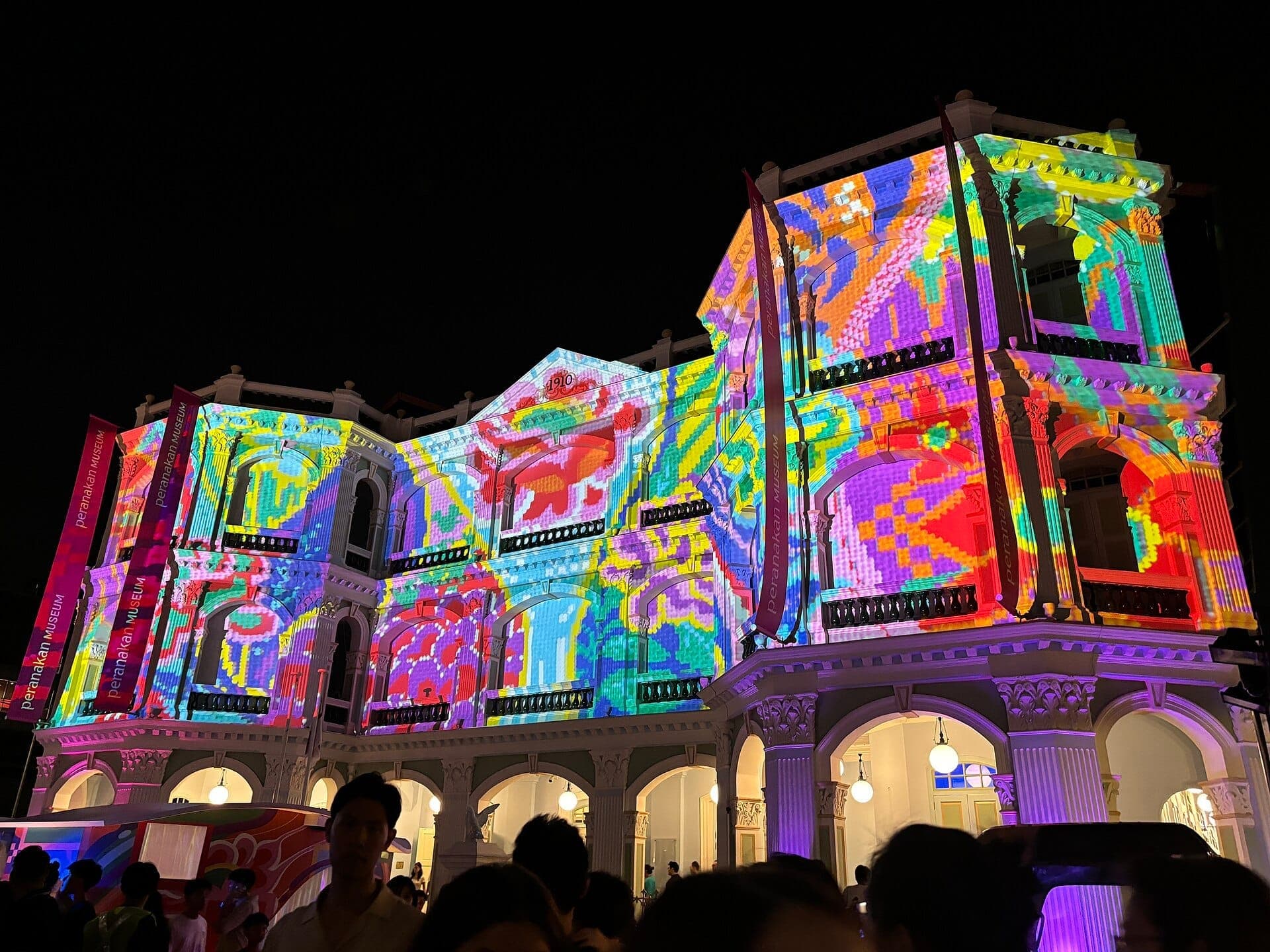 The Peranakan Museum building in Singapore illuminated at night with vibrant, colorful light projections on its colonial-era facade.