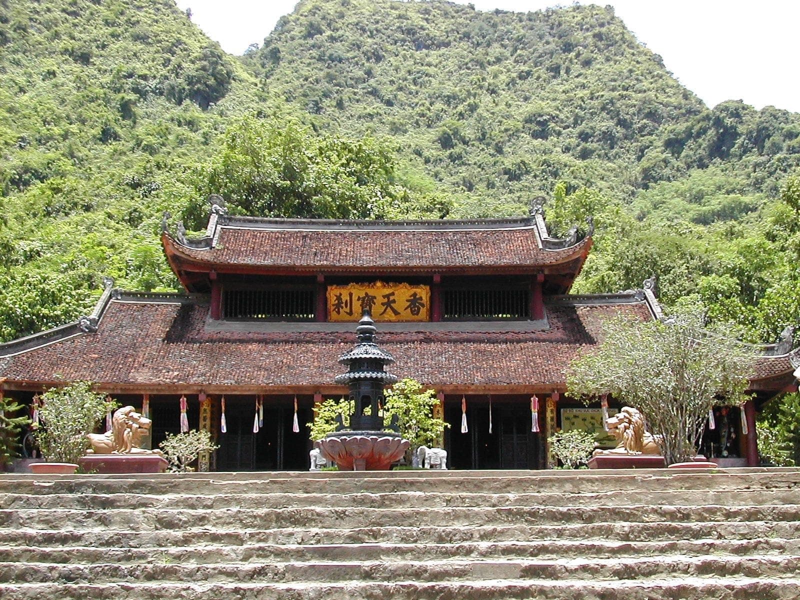 Lối vào chính của Chùa Hương với mái ngói đỏ truyền thống, bậc thềm đá, tượng sư tử và những ngọn núi xanh tươi tốt ở phía sau.