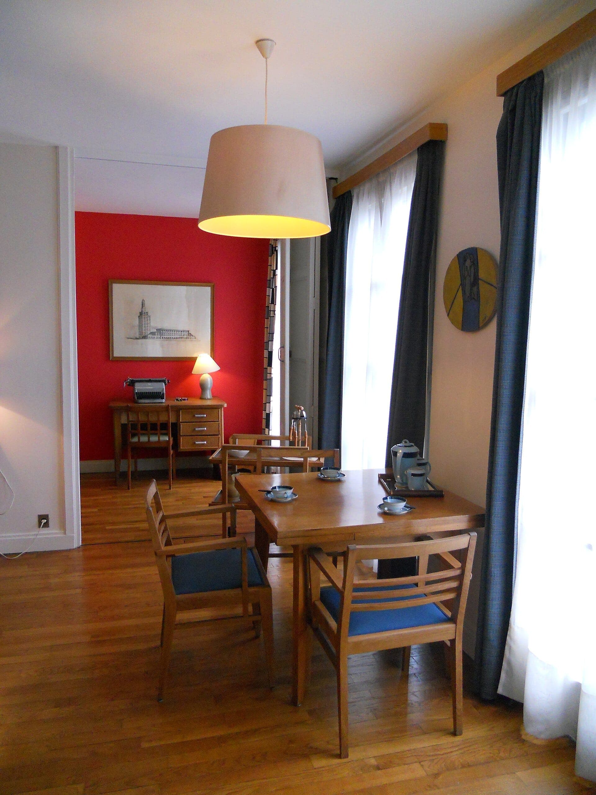 Phòng ăn hiện đại giữa thế kỷ bên trong Appartement Témoin Perret, với đồ nội thất bằng gỗ, ghế màu xanh, cửa sổ sáng và tường nhấn màu đỏ.
