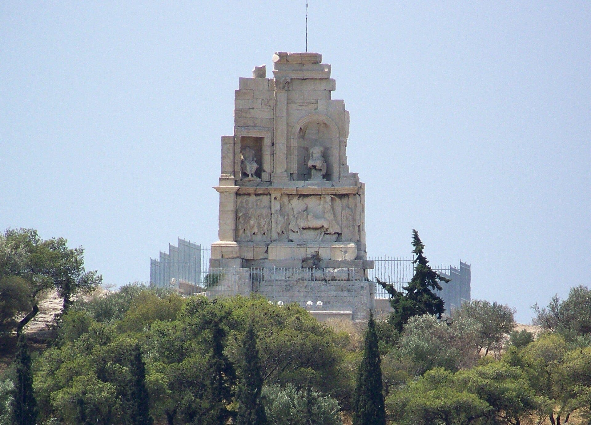 The Philopappos Monument rises above tree-covered slopes under a clear sky, its ancient marble details visible from a distance on the hilltop.