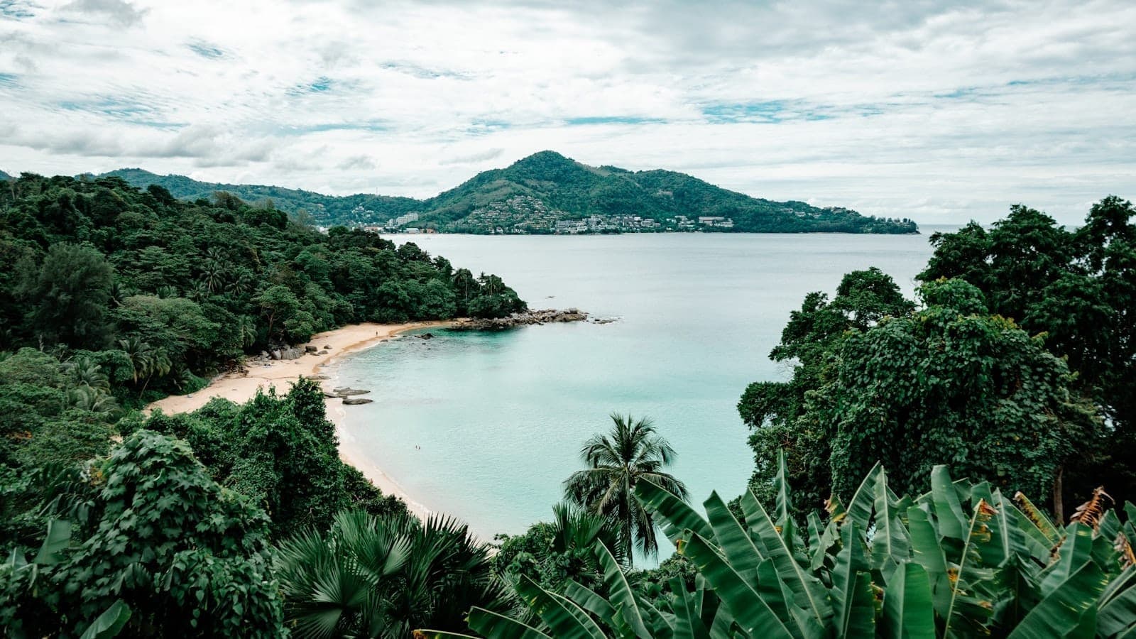 Toàn cảnh một bãi biển vắng vẻ ở Phuket được bao quanh bởi những ngọn đồi xanh mướt, biển xanh ngọc và dãy núi ở phía xa dưới bầu trời có mây trắng xen kẽ.