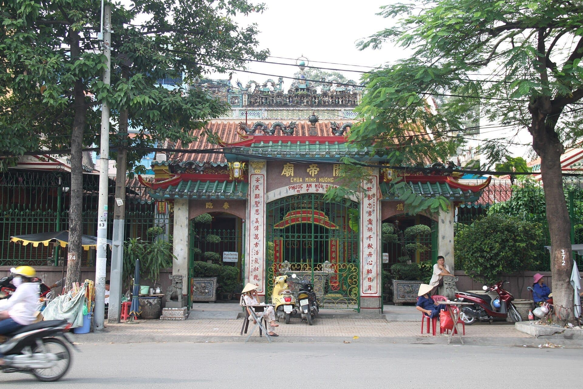 Mặt tiền chùa Phước An Hội Quán với mái ngói chạm khắc tinh xảo, cổng trang trí, cây cảnh trong chậu và người dân ngồi bên ngoài trên con phố tấp nập.